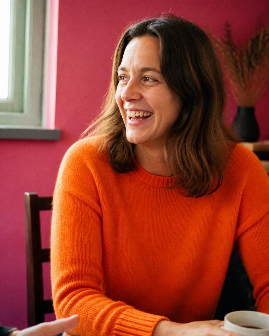 Woman in an orange jumper laughing in conversation against a pink wall